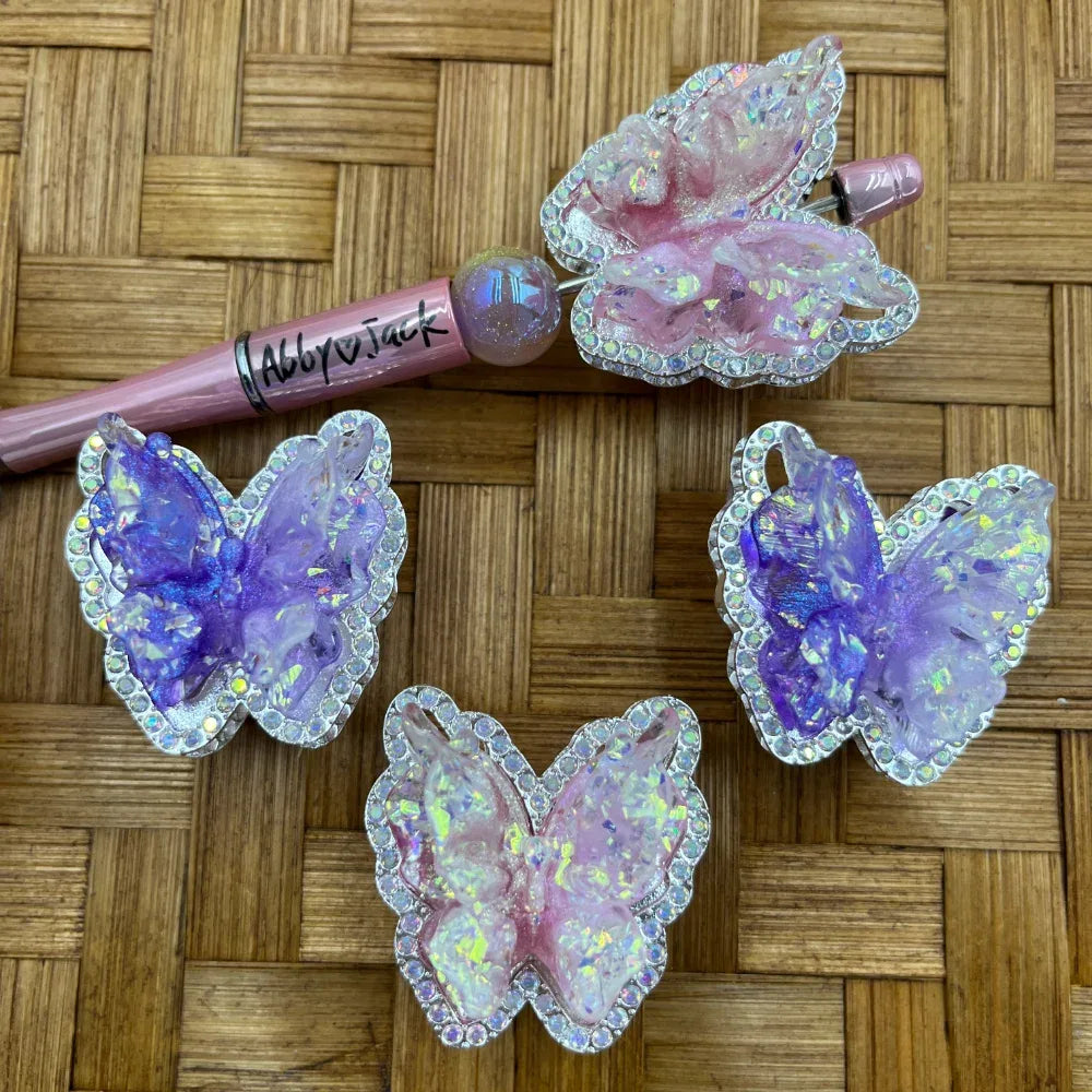 Four iridescent purple and pink resin butterfly beads with silver rhinestone borders displayed on a wicker surface next to a pink beadable pen with butterfly topper.