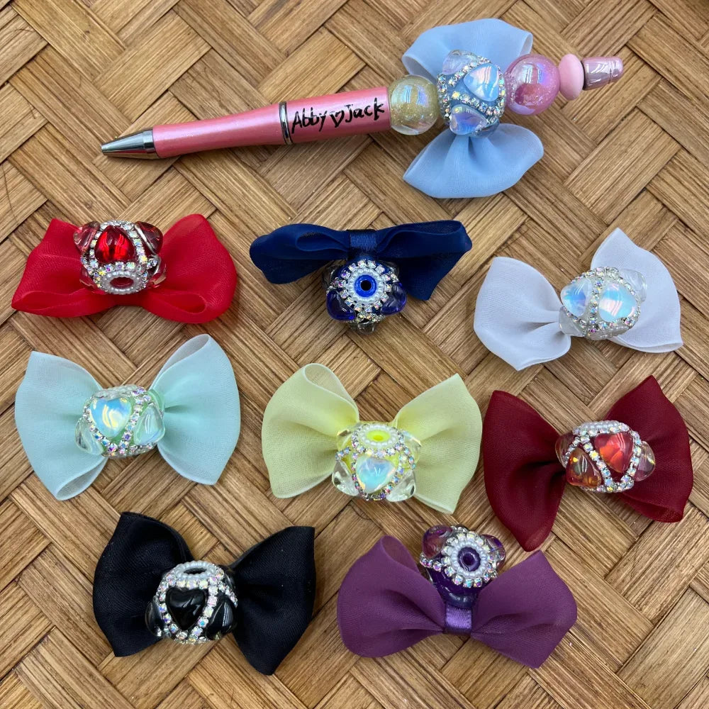 Assorted rhinestone bow fancy beads in red, navy, white, mint, yellow, burgundy, black, purple, and blue evil eye styles displayed with a decorative beadable pen on woven rattan background