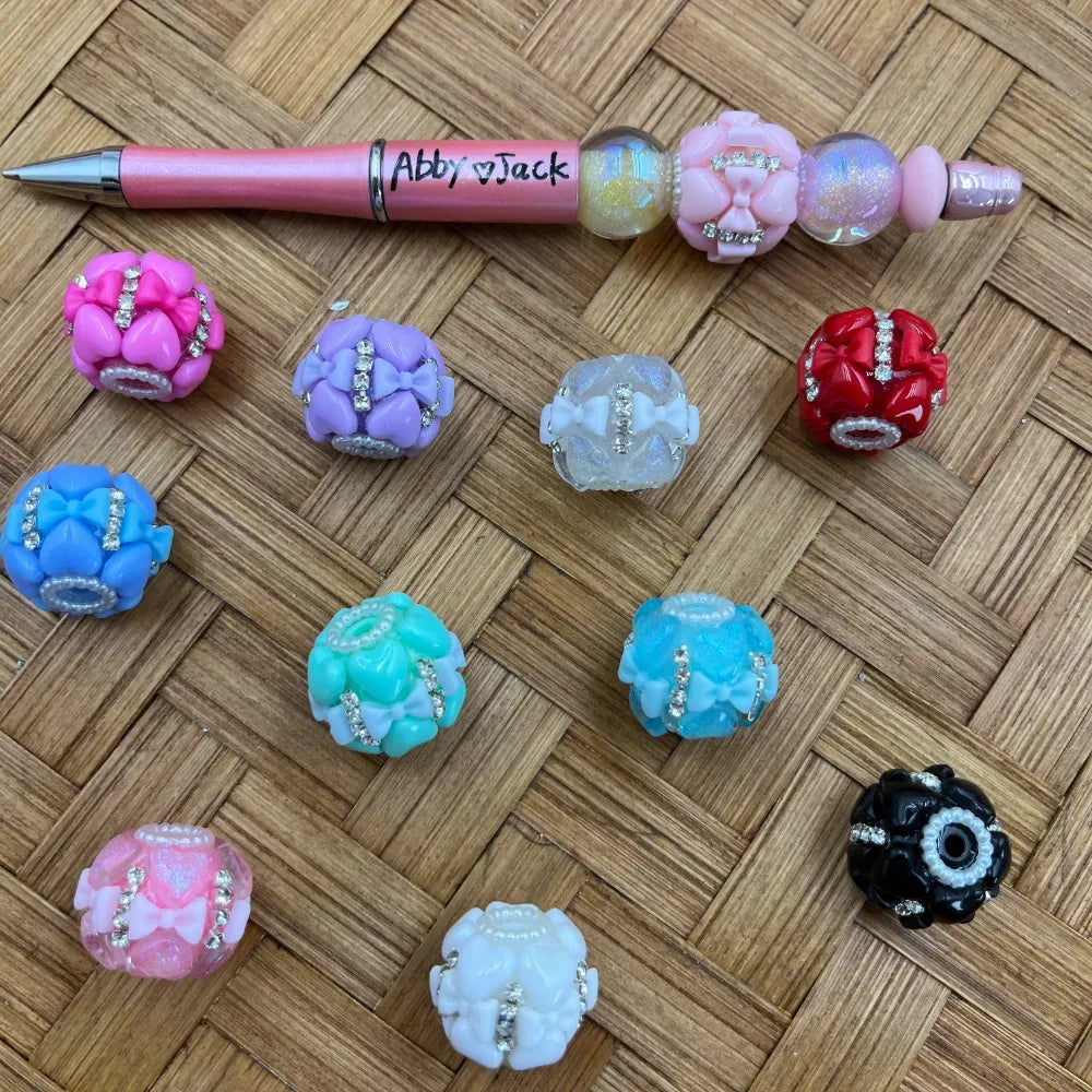 Assorted rhinestone bow fancy chunky beads in pink, purple, blue, teal, red, black, and white displayed on woven surface alongside a decorated beadable pen labeled Abby and Jack.