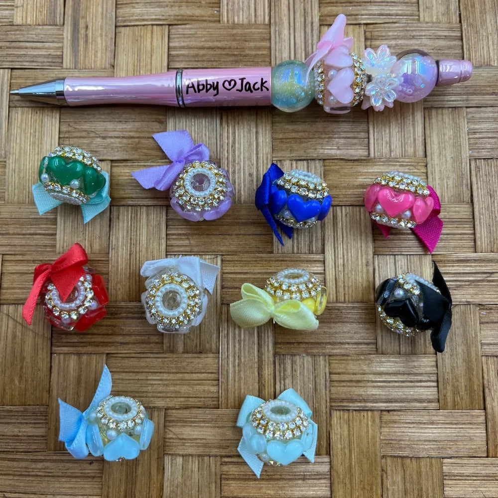 Assorted fancy beads decorated with rhinestone rims, ribbon bows, and pearl accents in multiple colors including red, blue, pink, black, yellow, and light blue, displayed next to a pink beadable pen labeled Abby and Jack on a wicker surface.