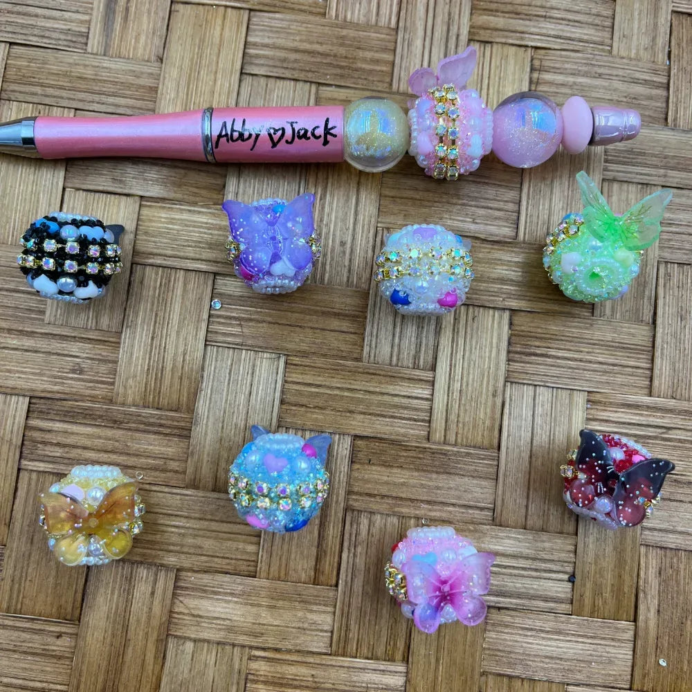 Assorted fancy decorative beads featuring rhinestone butterfly, purple cat, floral, and green dragonfly designs in pastel and dark colorways, displayed on woven bamboo surface alongside a pink beadable pen labeled Abby Jack.