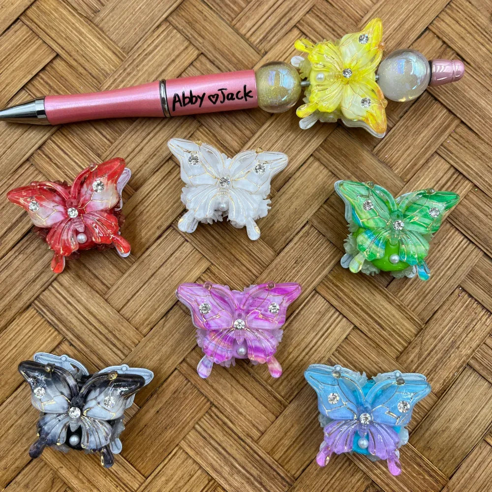 Assorted rhinestone butterfly resin fancy beads in red, white, yellow, green, purple, blue, and black displayed on woven surface next to a decorated beadable pen labeled Abby and Jack.