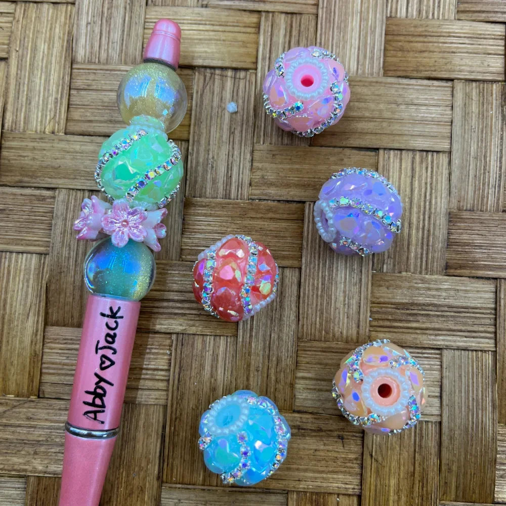 Colorful iridescent chunky resin beads with rhinestone bands in pastel pink, green, purple, coral, blue, and peach, shown alongside a decorated beadable pen on a wicker background.