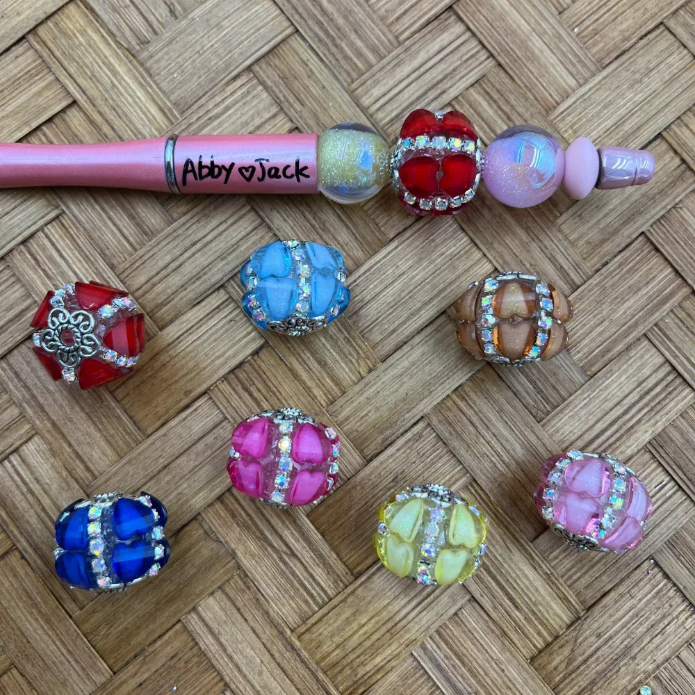 Assorted rhinestone crown chunky fancy beads in red, blue, pink, yellow, teal, brown, and light pink colors displayed on a woven surface alongside a pink beadable pen labeled Abby Jack, showing the beads threaded on the pen.