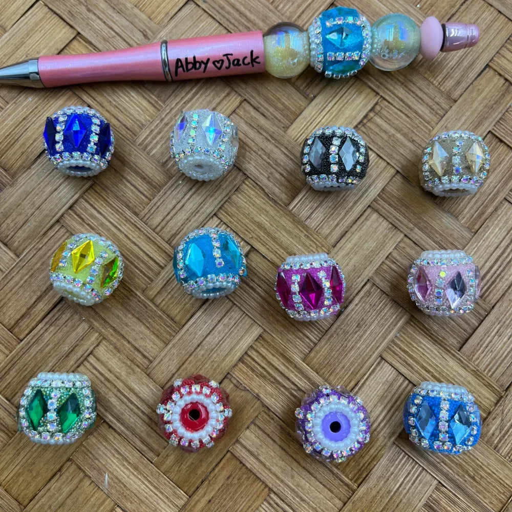Assorted rhinestone crown fancy beads with colorful gemstone diamond centers and pearl or crystal-rimmed edges in blue, white, black, gold, yellow, teal, pink, purple, green, and red, displayed on a wicker surface next to a beaded pen labeled Abby Jack.