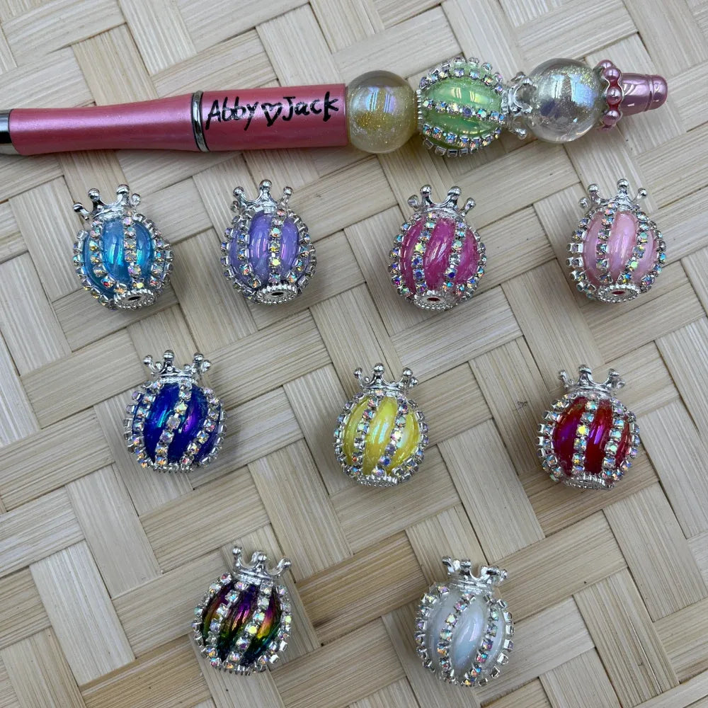 Nine rhinestone-bordered crown ladybug fancy beads in assorted colors including blue, purple, pink, yellow, red, rainbow, and white, displayed on a woven mat next to a beadable pen