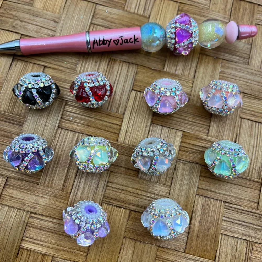 Assorted rhinestone-encrusted crystal fancy beads in black, red, purple, green, pink, and iridescent AB colors displayed on a woven surface next to a beaded pen labeled Abby Jack.