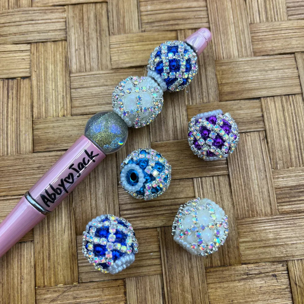 Assorted rhinestone crystal fancy beads in blue, purple, and white with AB sparkle finish displayed next to a pink beadable pen on a wicker surface