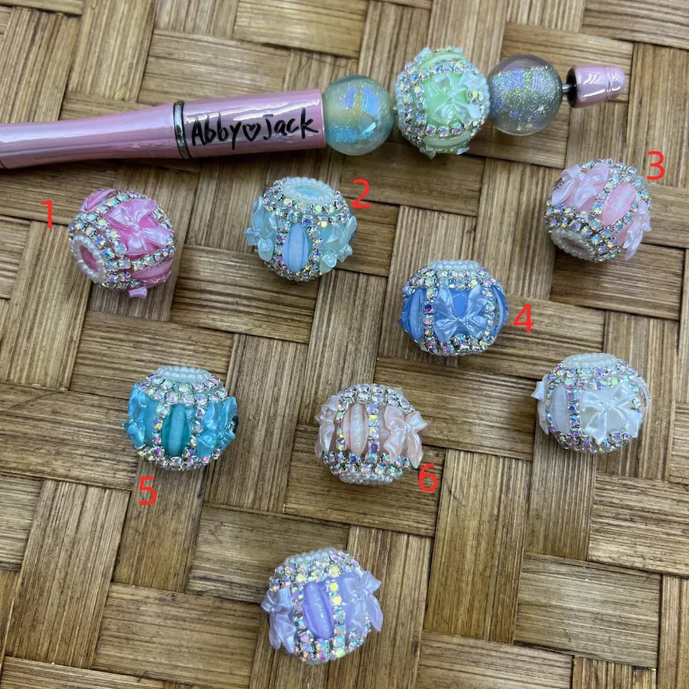 Set of 7 rhinestone-encrusted fancy beads in pastel pink, blue, green, and white with floral bow and cat-eye accents, numbered 1-6 plus an assembled beadable pen on wicker surface.