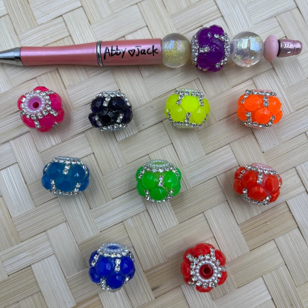 Assorted colorful rhinestone crystal cluster fancy beads in pink, black, yellow, orange, blue, green, and red displayed on a woven surface next to a pink beadable pen labeled Abby Jack.