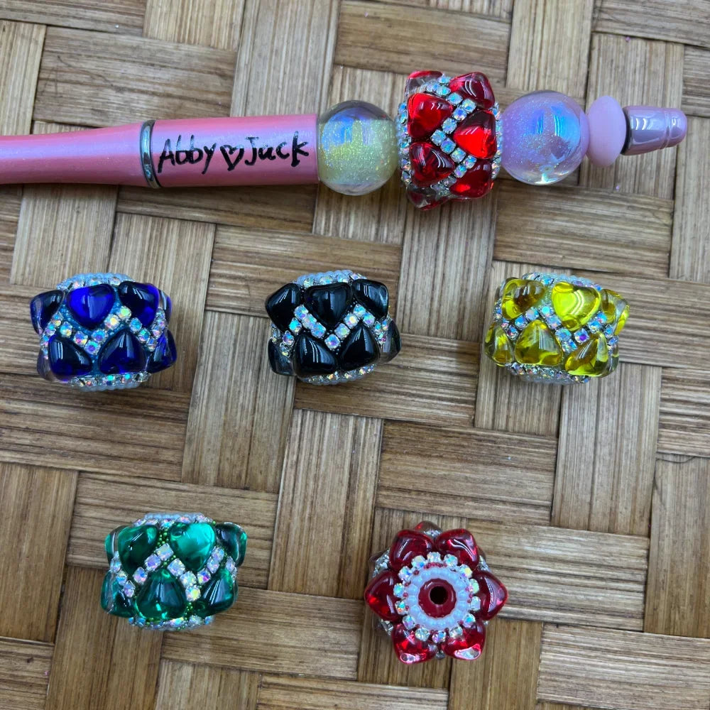 Assorted rhinestone-encrusted crystal fancy barrel beads in red, blue, black, yellow, green, and iridescent colors displayed on a woven surface next to a pink beadable pen branded Abby Jack.