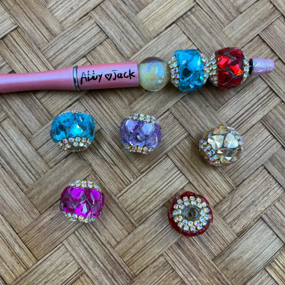 Assorted rhinestone crystal fancy beads in teal, red, purple, pink, and clear colors shown on a woven surface next to a pink beadable pen labeled Abby and Jack.
