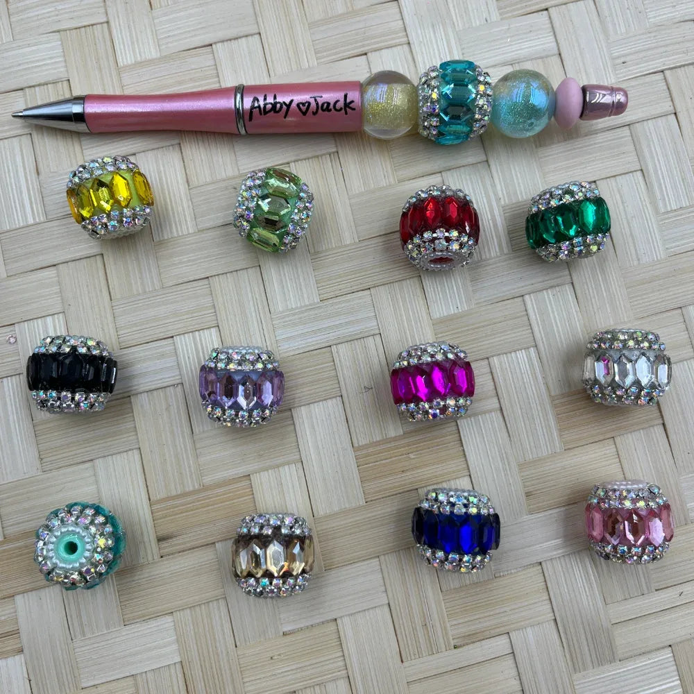 Assorted rhinestone-encrusted crystal fancy beads in multiple colors including yellow, green, red, teal, black, lavender, magenta, clear, teal, champagne, navy, and pink, displayed on a woven surface next to a decorative beaded pen labeled Abby Jack.