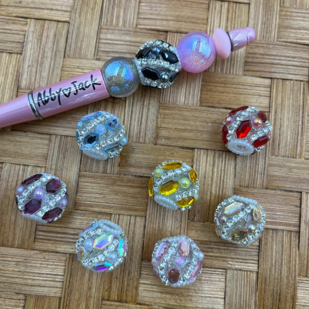 Assorted rhinestone crystal fancy beads in multiple colors including black, red, yellow, pink, purple, blue, and iridescent AB, displayed next to a pink beadable pen on a wicker surface.