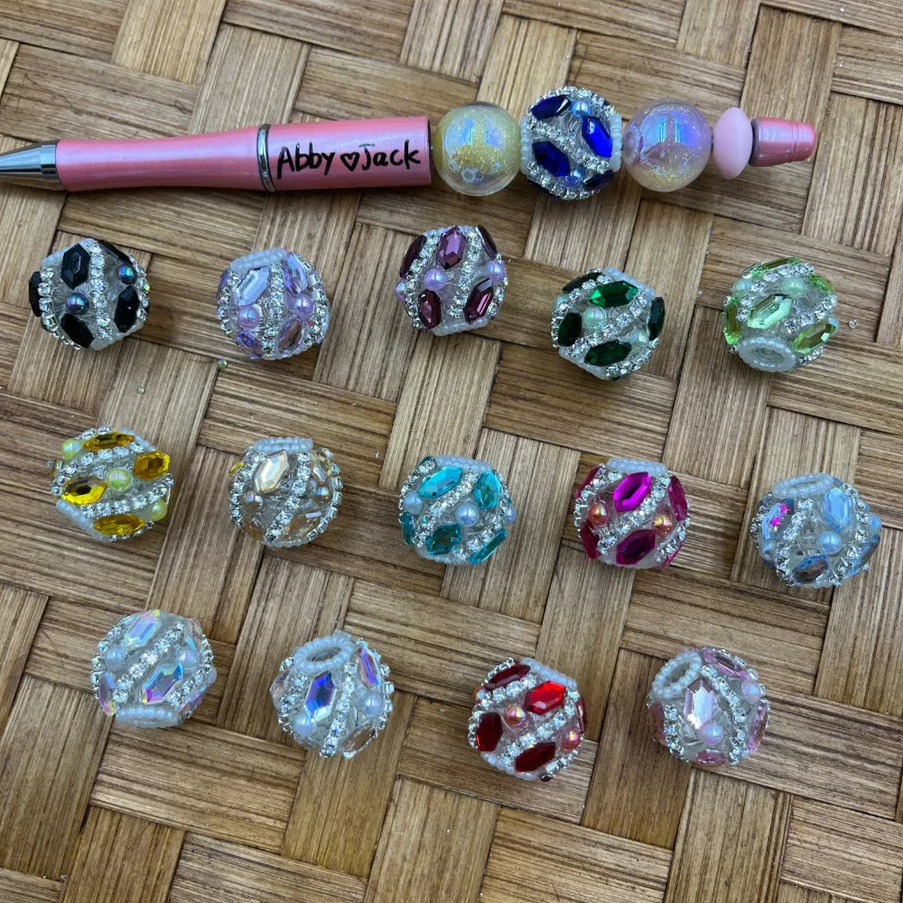 Assorted rhinestone crystal fancy chunky beads with colorful gemstone accents in black, purple, red, green, yellow, teal, pink, and iridescent finishes, displayed next to a pink beadable pen on a woven surface.
