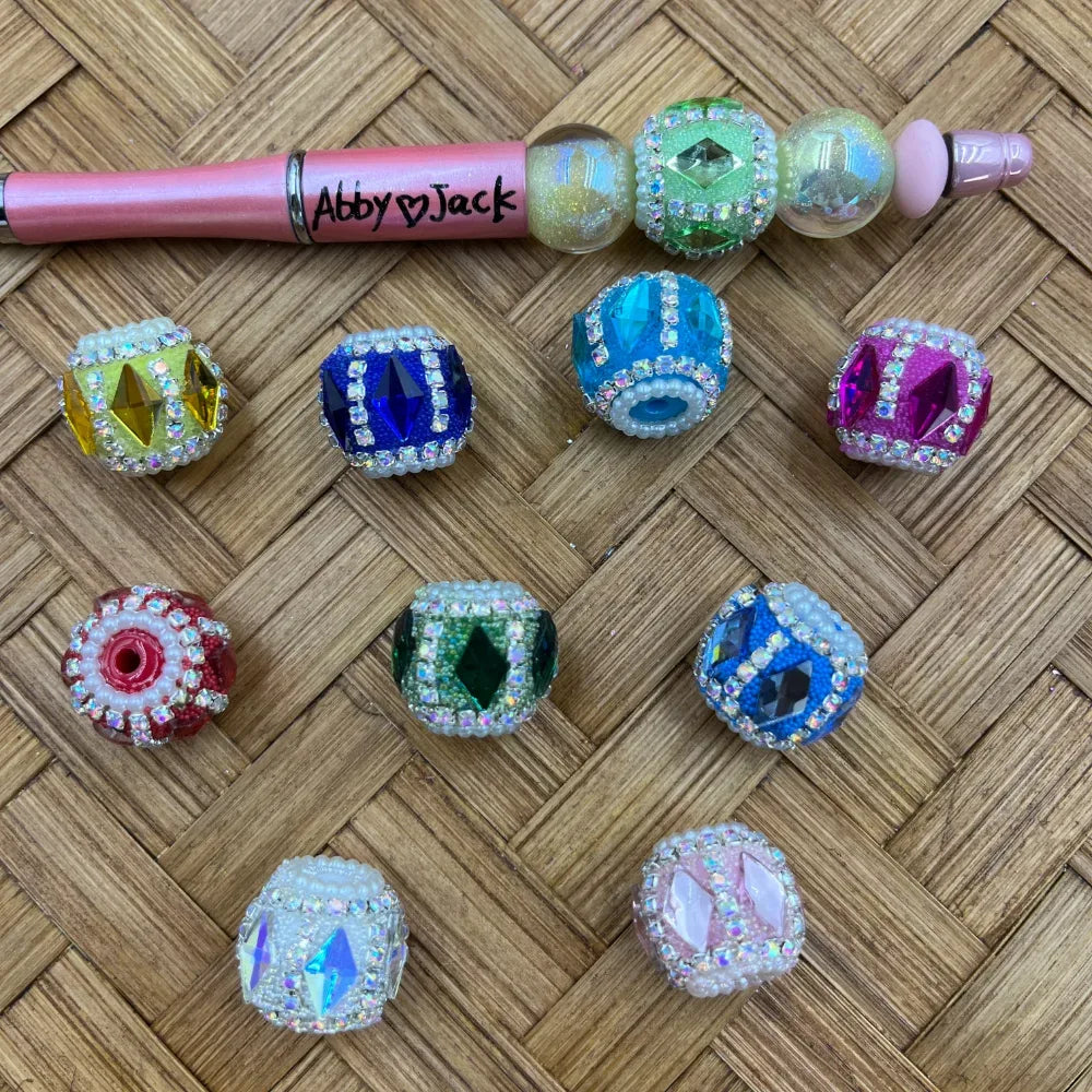 Assorted rhinestone crystal gem fancy beads in yellow, blue, teal, pink, red, green, white, and light blue with pearl and AB crystal accents, displayed on woven rattan surface alongside a pink beadable pen labeled Abby Jack.
