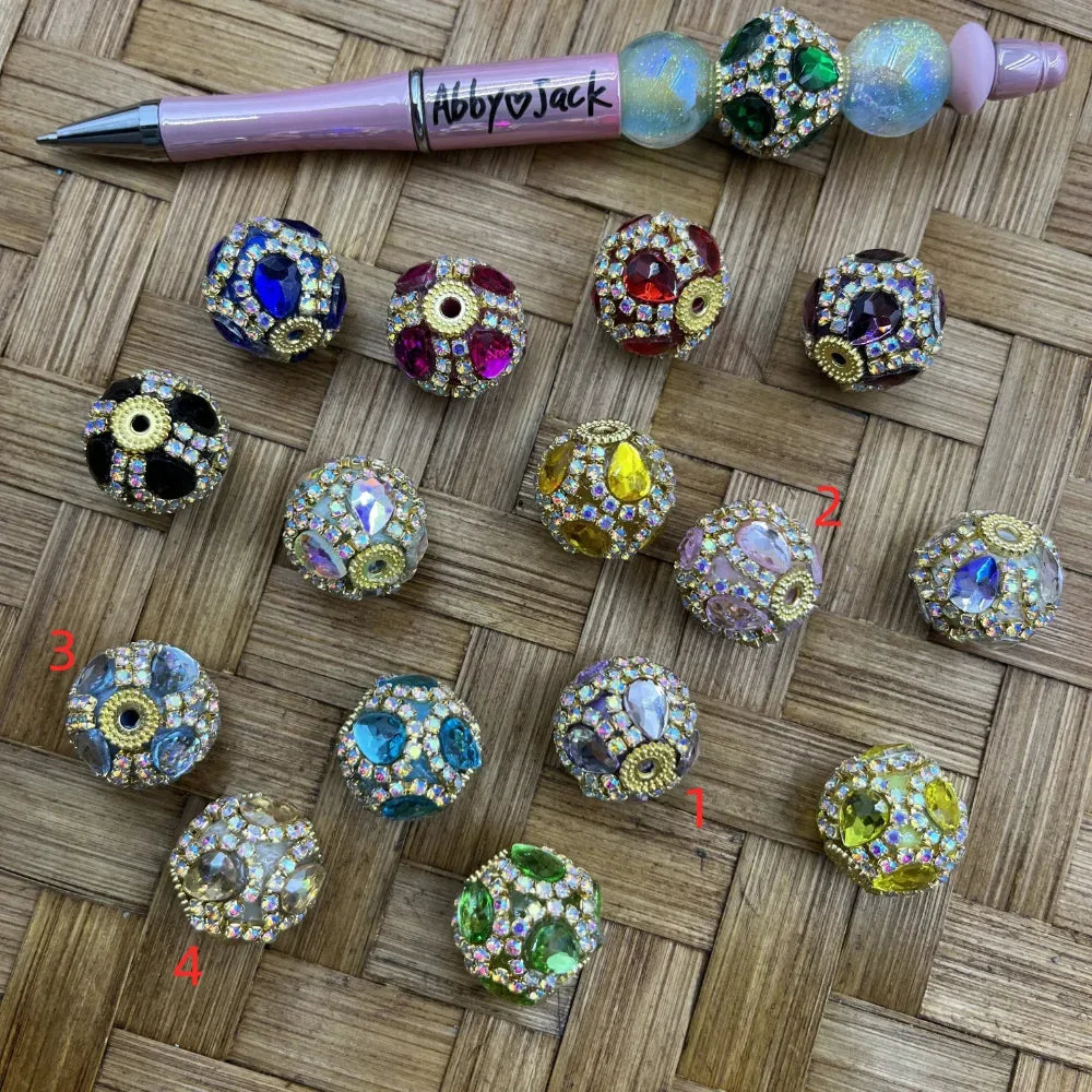 Assorted rhinestone crystal gem fancy beads in multiple colors including blue, red, pink, purple, teal, yellow, green and clear AB, set in gold metal with large center stones and micro pave rhinestone accents, displayed on woven surface next to a beadable pen.