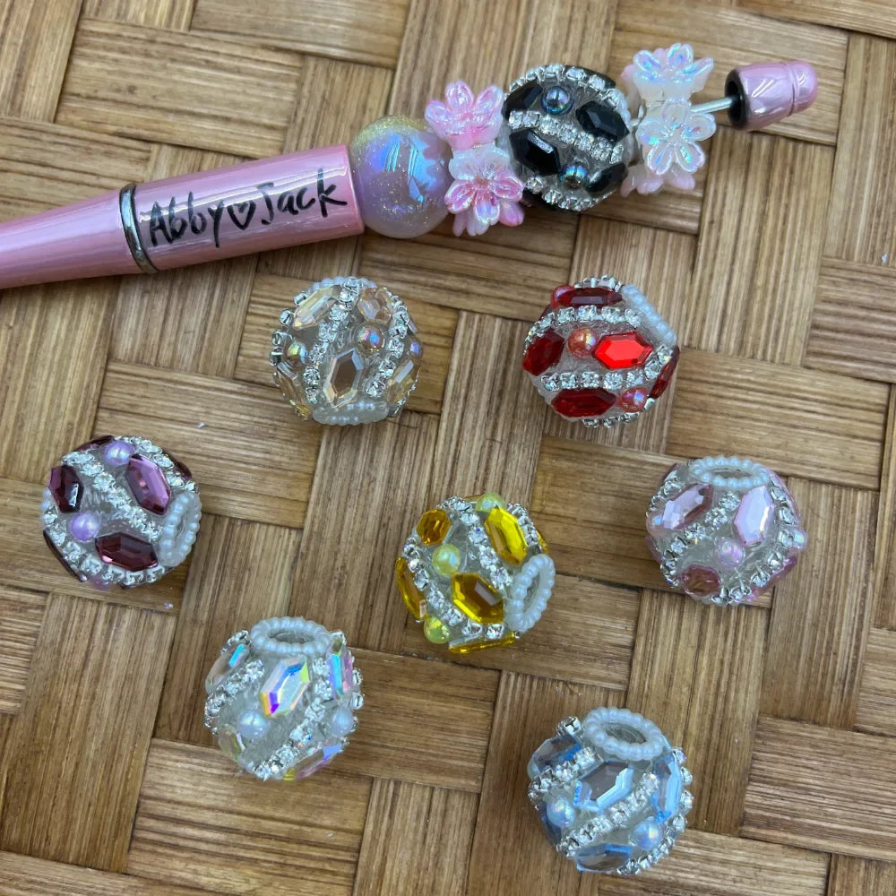 Assorted rhinestone crystal gem fancy beads in red, purple, yellow, pink, clear, and black colorways displayed on woven bamboo surface next to a decorated beadable pen labeled Abby and Jack