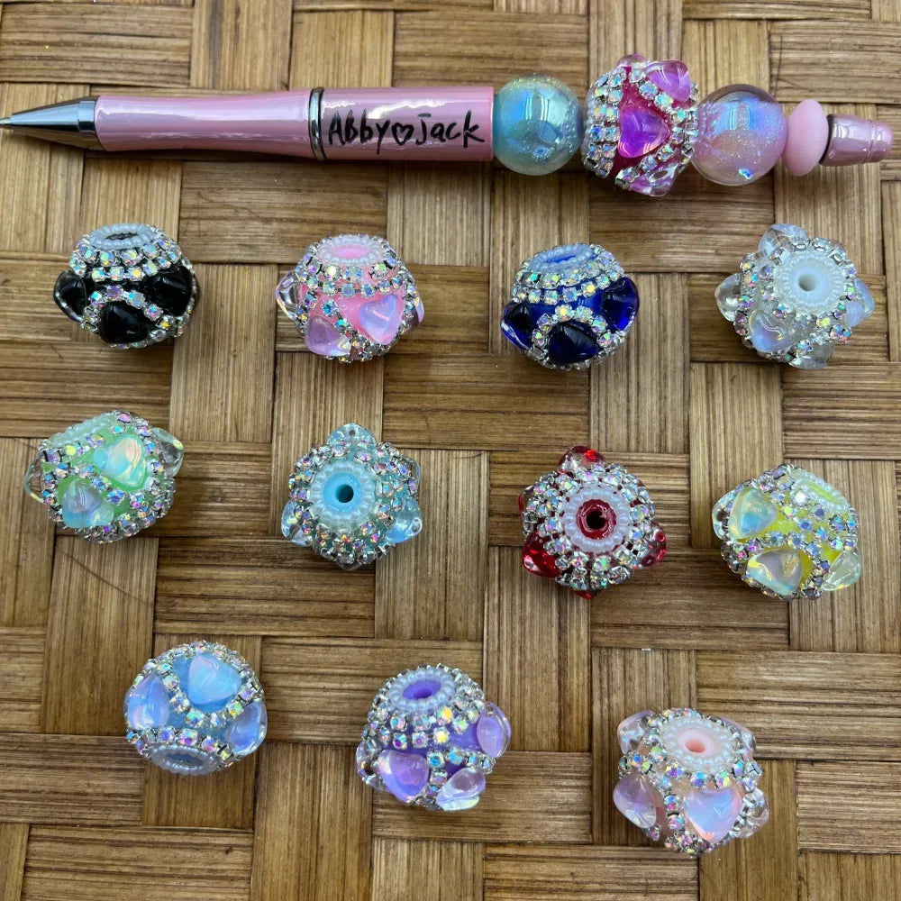 Assorted rhinestone-encrusted crystal heart fancy beads in multiple colors including black, pink, blue, red, purple, and iridescent, displayed on a woven surface next to a pink beadable pen labeled Abby Jack.