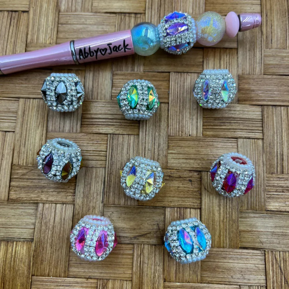 Assorted rhinestone crystal fancy beads with colorful marquise and faceted gemstone centers in pink, green, blue, purple, and aurora borealis finishes, displayed on woven bamboo surface next to a decorated beadable pen.