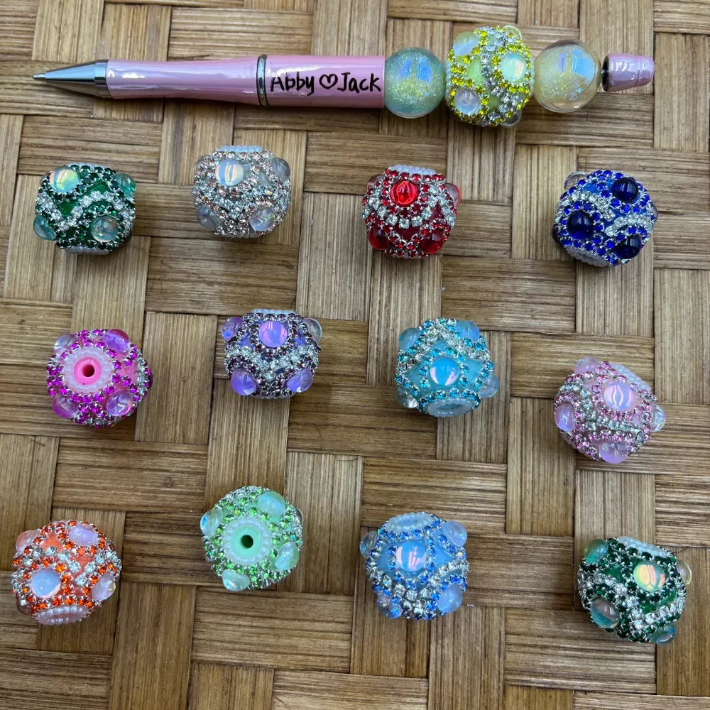 Assorted rhinestone crystal fancy beads in multiple colors including green, silver, red, blue, pink, purple, teal, orange, and mint, displayed on a woven bamboo surface alongside a pink beadable pen labeled Abby and Jack, for use in DIY jewelry making, keychains, and badge reels.