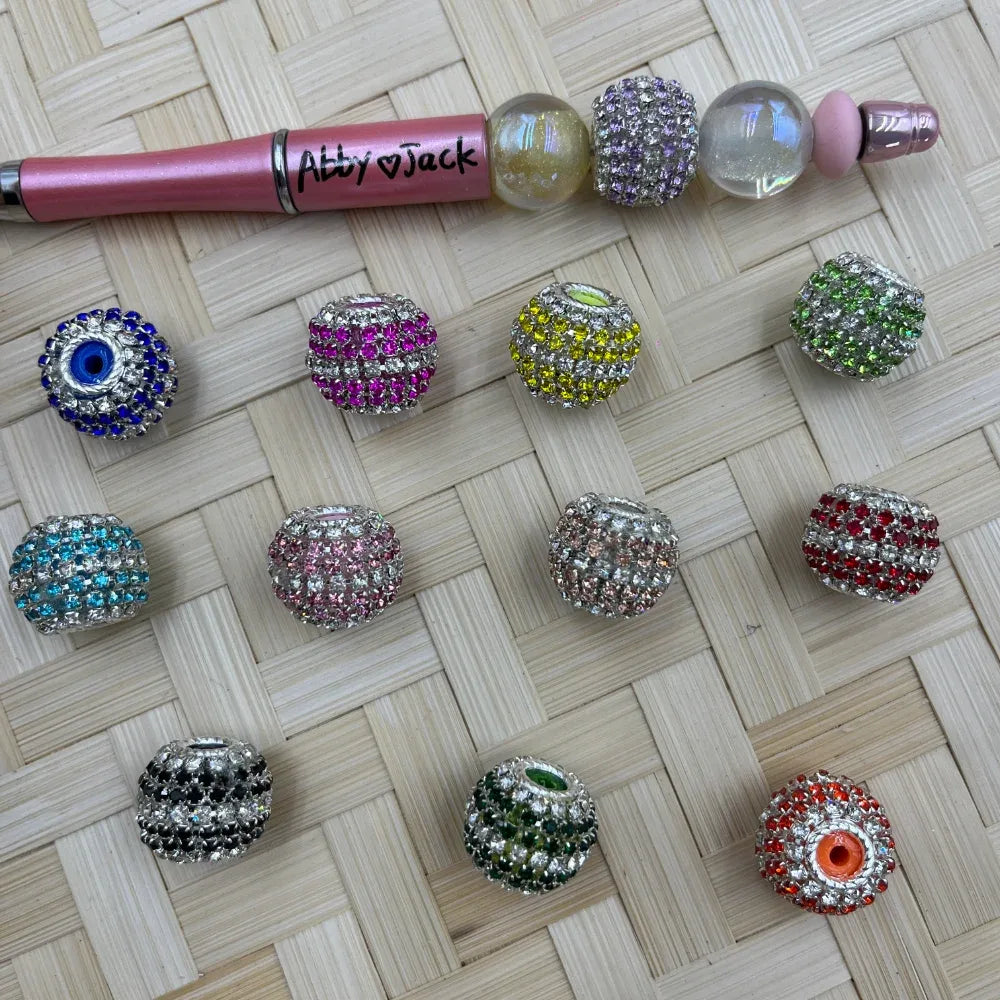 Assorted rhinestone crystal pave fancy beads in 12 colors including blue evil eye, hot pink, yellow, green, teal, light pink, peach, red, black, dark green, and orange evil eye displayed on woven bamboo surface next to a beadable pen labeled Abby Jack.