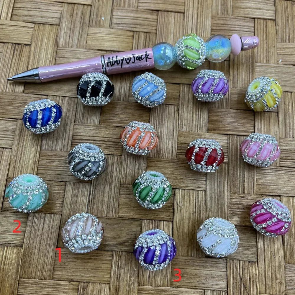Assorted rhinestone-encrusted crystal stripe fancy beads in multiple colors including blue, black, green, orange, red, purple, pink, and yellow, displayed on a woven surface next to a beadable pen.