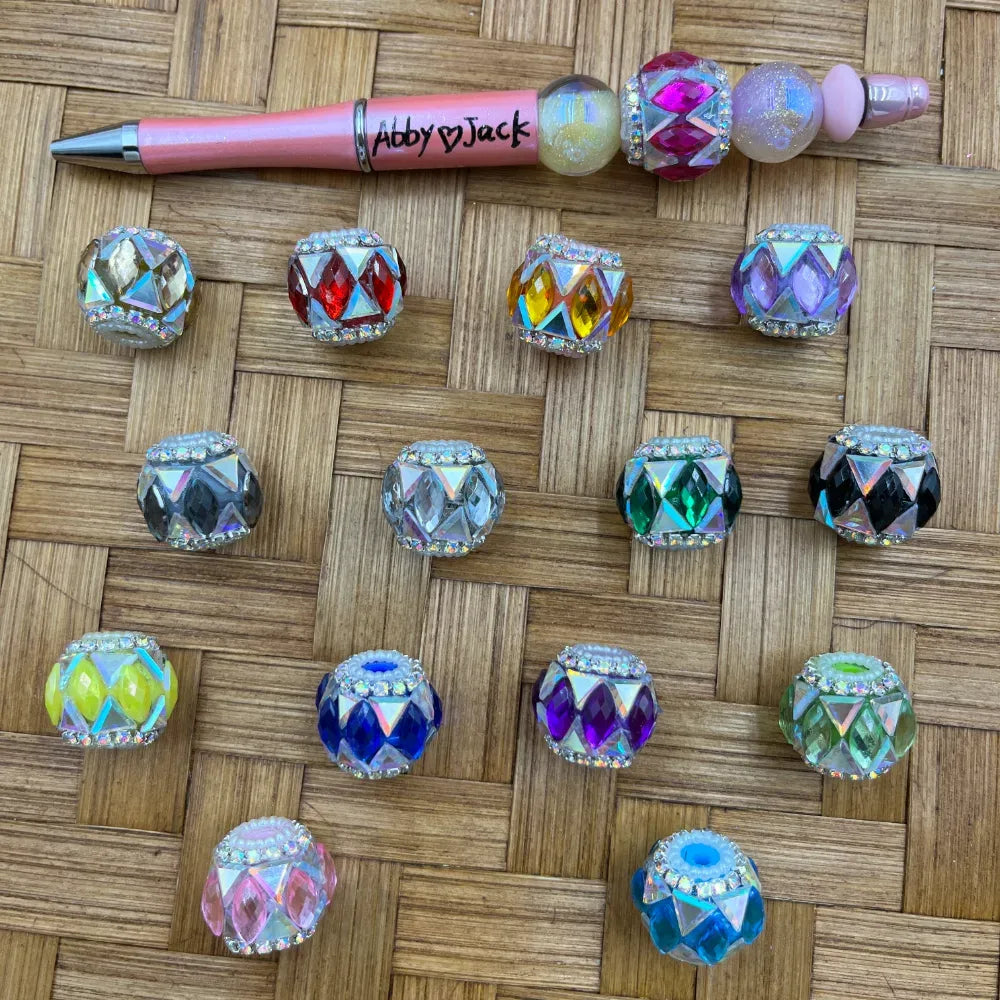 Assorted rhinestone-rimmed diamond-cut crystal fancy beads in multiple colors including red, purple, green, blue, black, pink, and yellow, displayed on a woven surface next to a decorated beadable pen labeled Abby Jack.