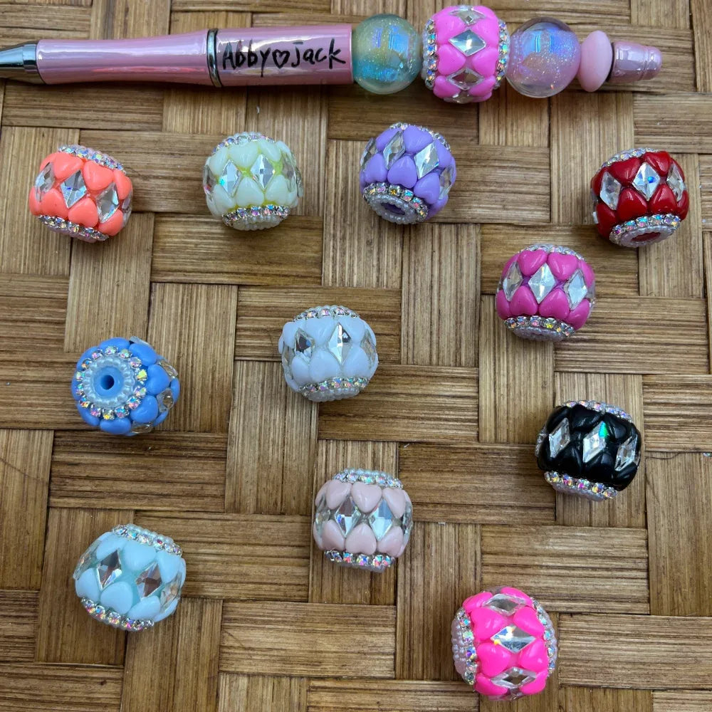 Assorted colorful rhinestone diamond fancy beads in pink, orange, purple, blue, red, black, white, and hot pink with crystal embellishments, displayed on a woven surface next to a decorated beadable pen labeled Abby Jack.