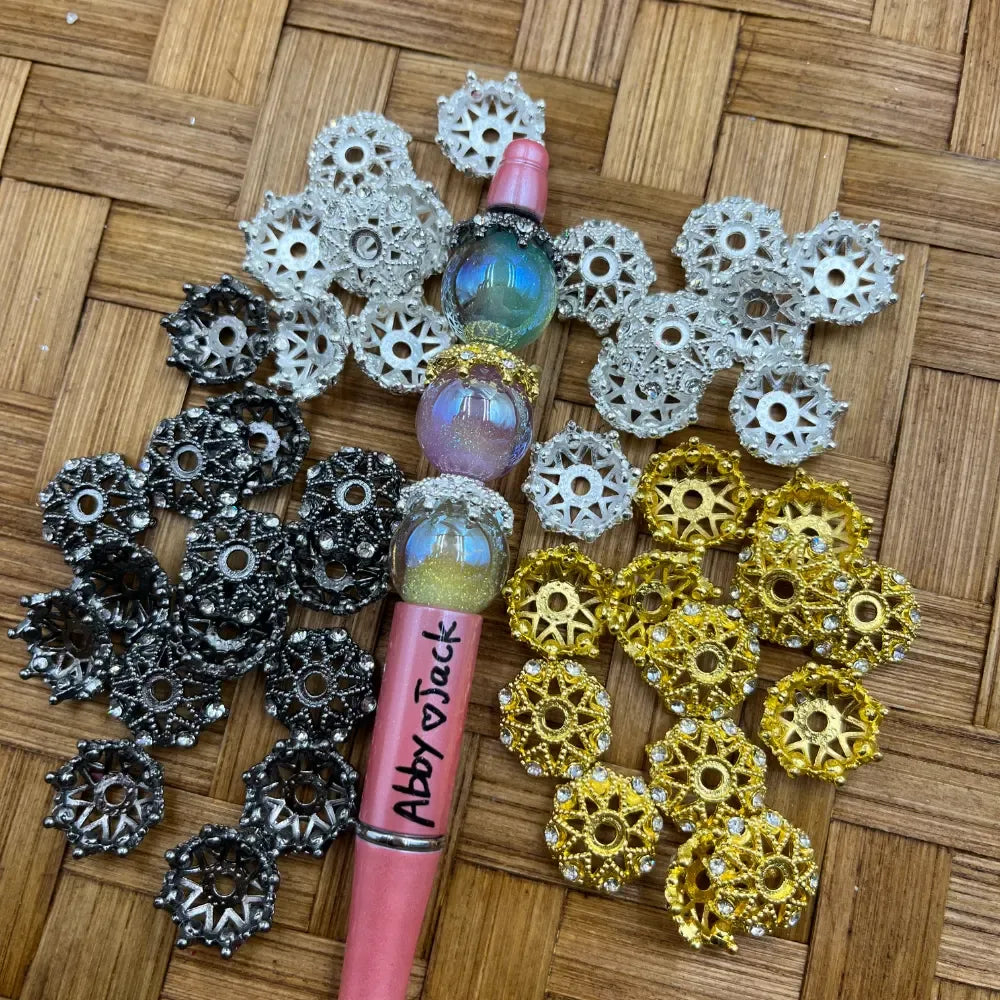 Assorted rhinestone filigree metal bead caps in gold, silver, and gunmetal black colors displayed around a pink beadable pen with iridescent glitter beads on a wicker background.