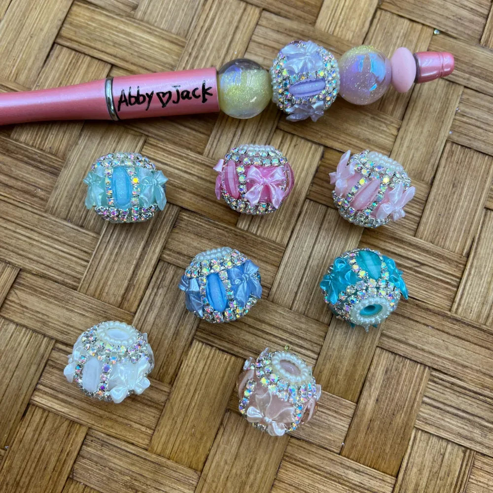 Assorted pastel rhinestone floral bow chunky fancy beads in blue, pink, teal, white, and lavender displayed on a woven surface next to a decorated beadable pen labeled Abby Jack.