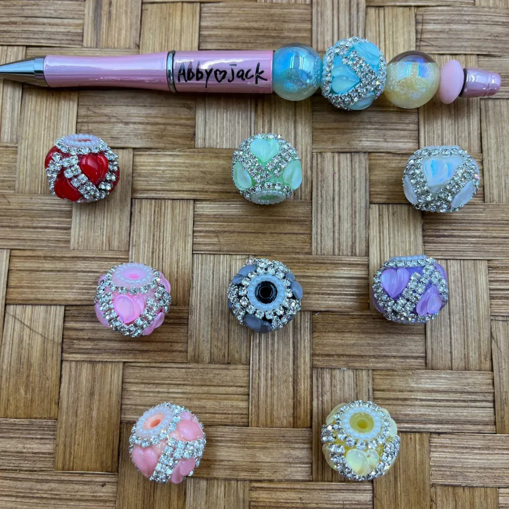 Assorted rhinestone-encrusted fancy beads with floral heart accents in red, pink, green, blue, purple, gray, and yellow, displayed next to a pink beadable pen on a wicker background.