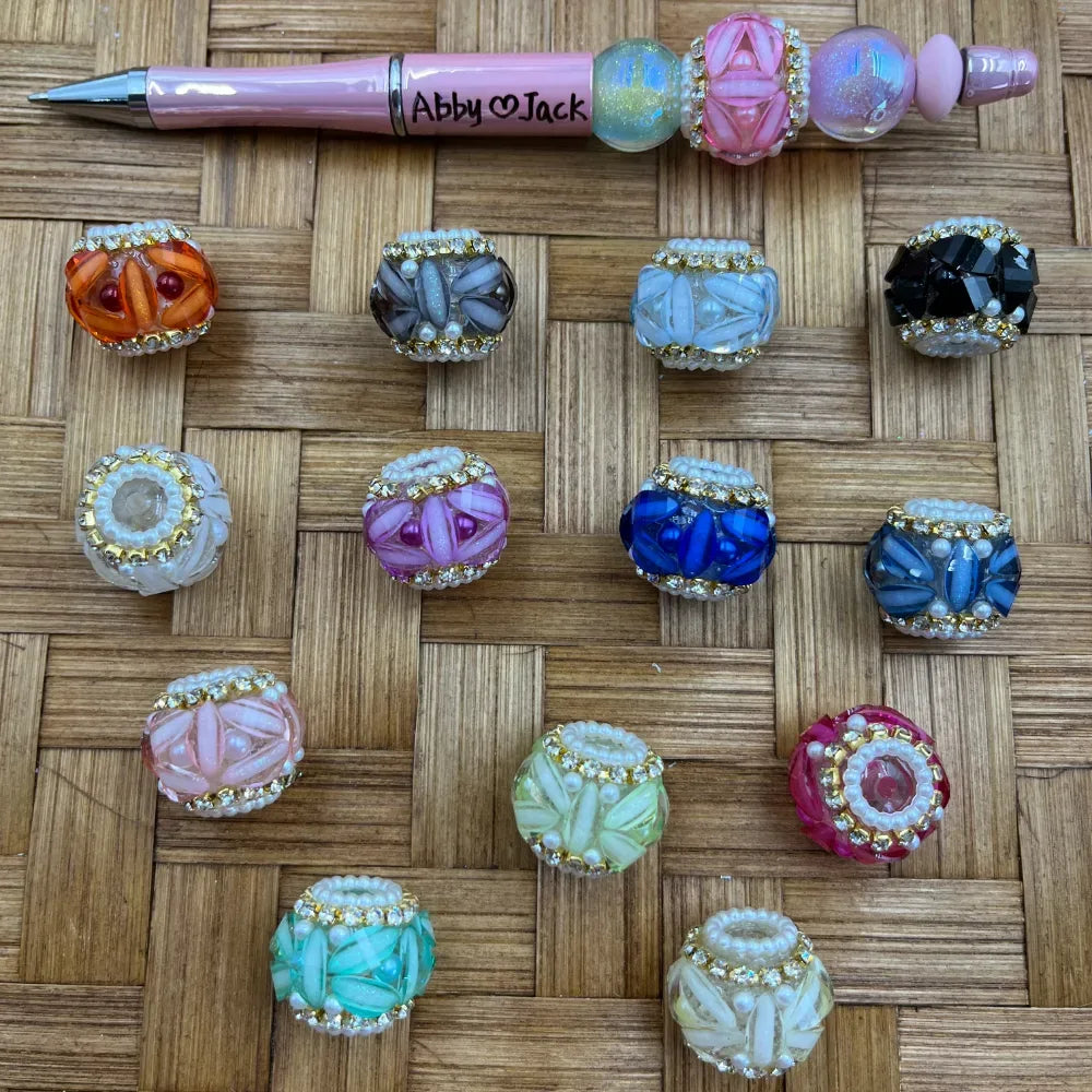 Assorted rhinestone-trimmed floral petal fancy beads in multiple colors including pink, blue, orange, black, teal, and mint, displayed on a woven surface next to a beaded pen labeled Abby and Jack.