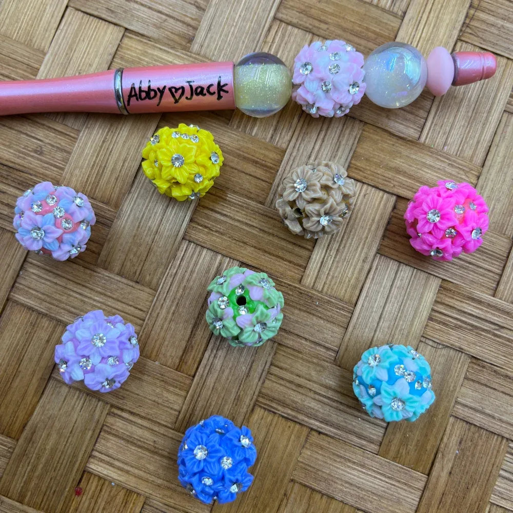 Assorted color rhinestone flower ball fancy beads in yellow, pink, blue, lavender, green, tan, and teal displayed on a wicker surface next to a beadable pen labeled Abby Jack.