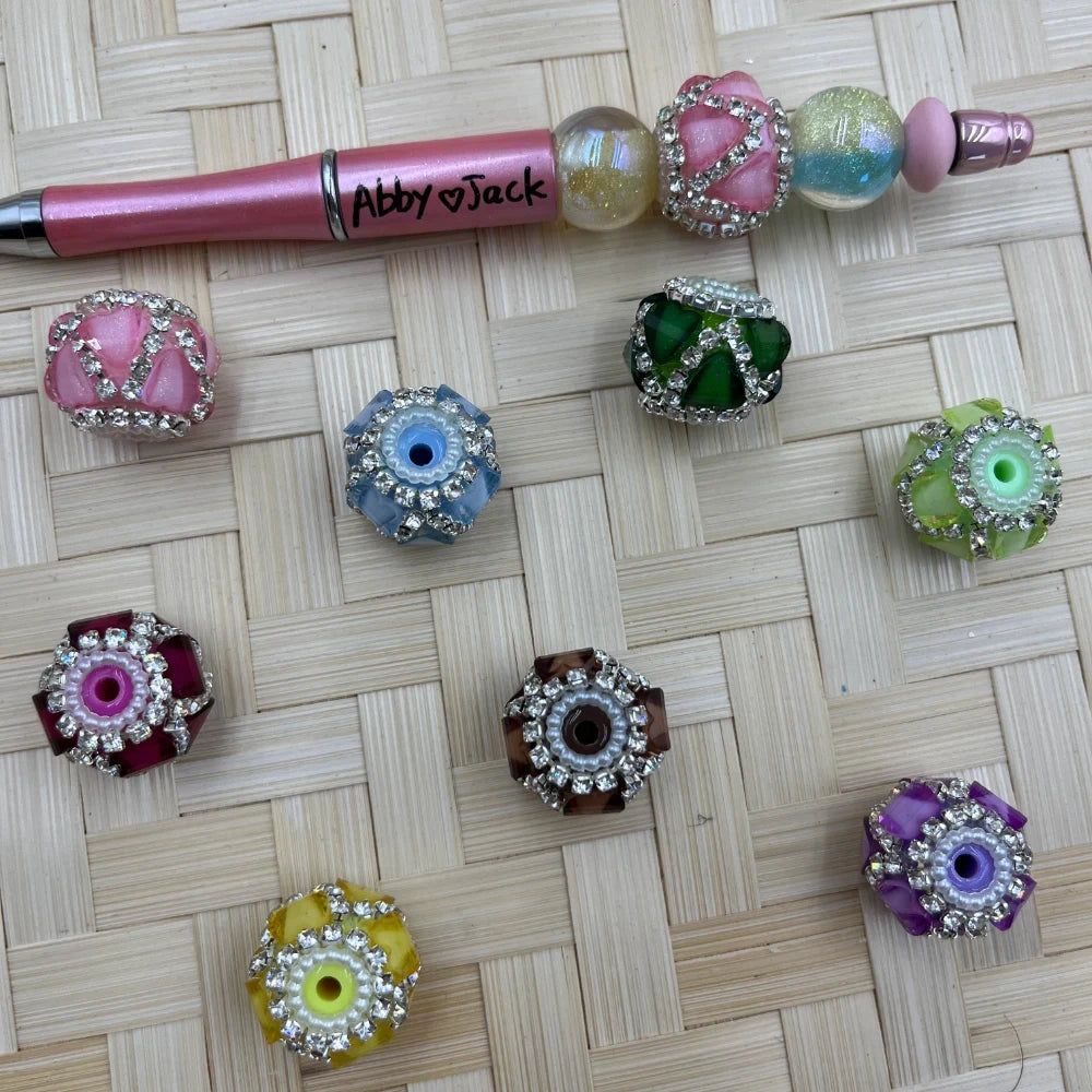 Assorted rhinestone-trimmed flower petal fancy beads in pink, blue, green, purple, yellow, burgundy, and brown next to a pink beadable pen labeled Abby Jack on a woven bamboo surface.