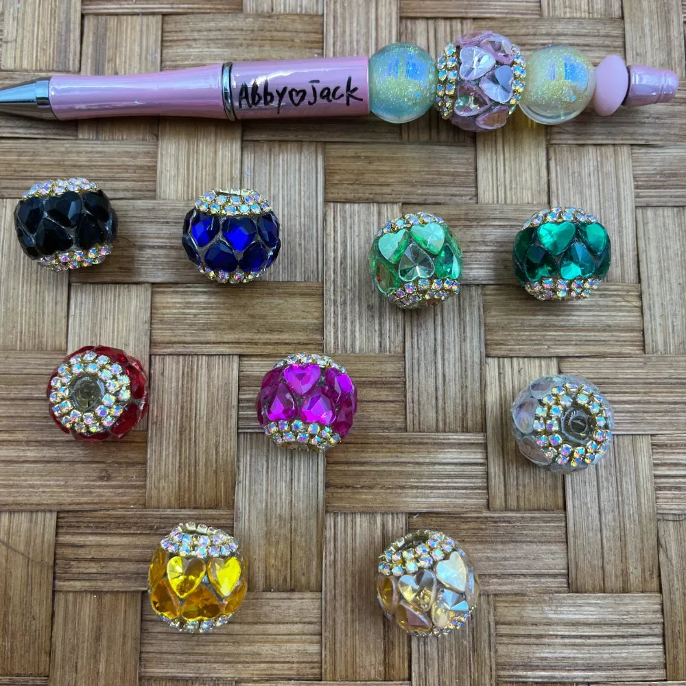 Nine rhinestone-rimmed heart crystal fancy beads in assorted colors including black, navy blue, light green, emerald green, red, hot pink, clear/iridescent, yellow, and white AB, displayed on a woven bamboo surface next to a pink beadable pen labeled Abby and Jack.
