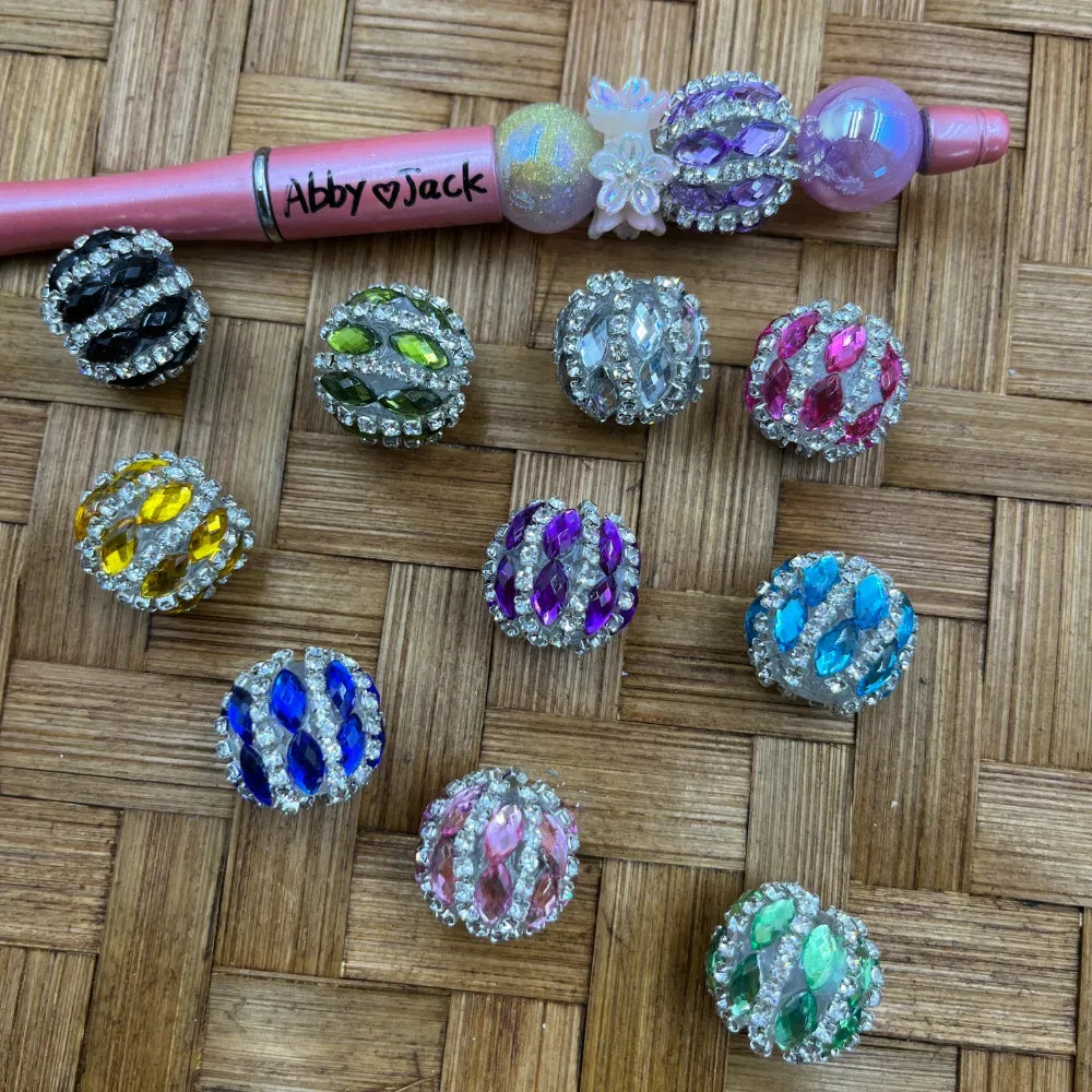 Assorted rhinestone-encrusted marquise crystal fancy beads in multiple colors including black, green, clear, pink, yellow, purple, blue, teal, light pink, and mint, displayed on a woven surface next to a decorated beadable pen.
