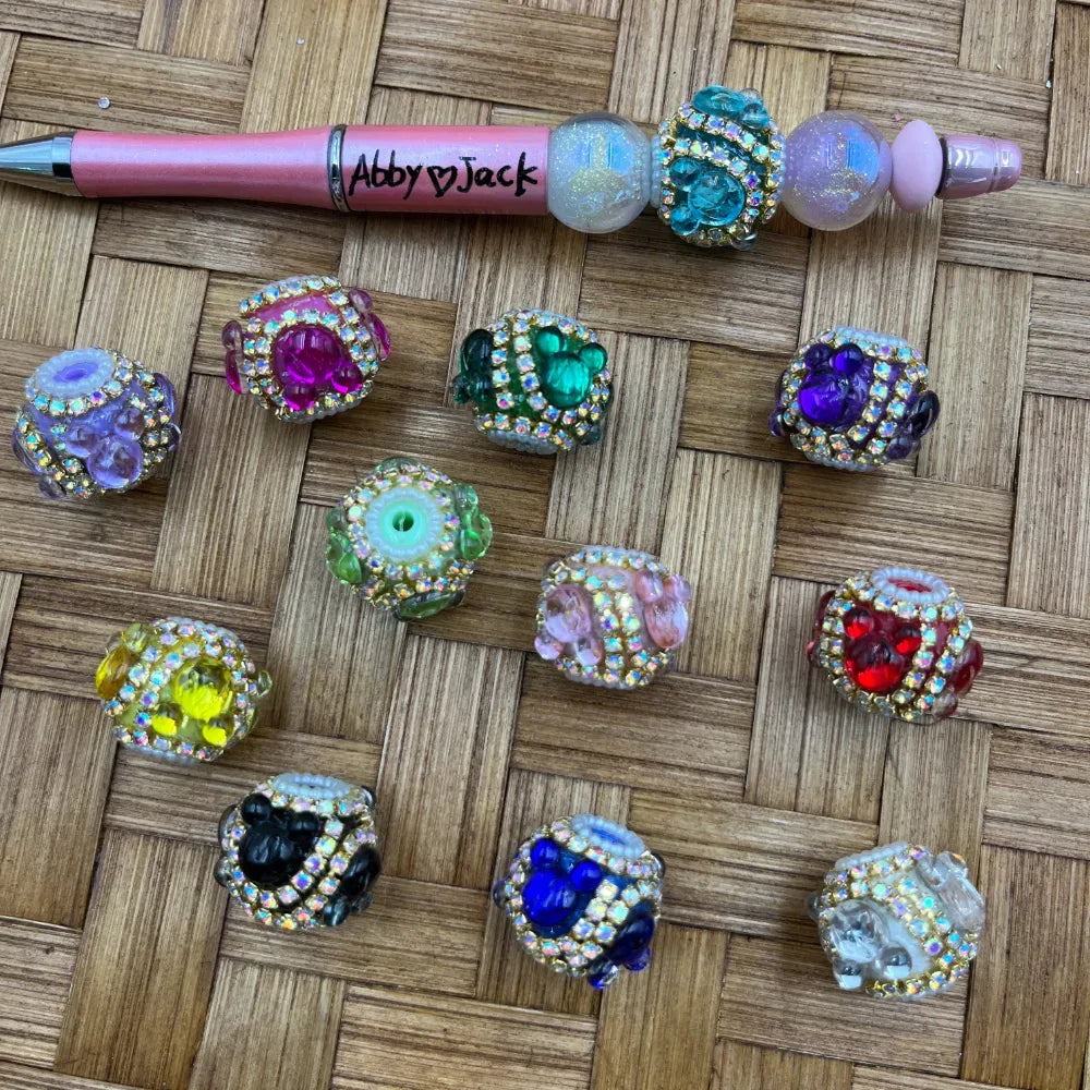 Assorted rhinestone-rimmed Mickey Mouse shaped fancy cube beads in multiple colors including pink, green, purple, yellow, red, blue, black, and clear, displayed on a woven surface alongside a pink beadable pen labeled Abby Jack.