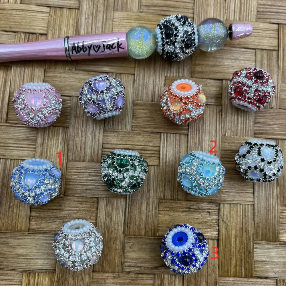 Assorted rhinestone pave crystal fancy beads in pink, purple, orange, red, blue, green, teal, black and white colorways displayed on woven surface alongside a beadable pen labeled Abby and Jack