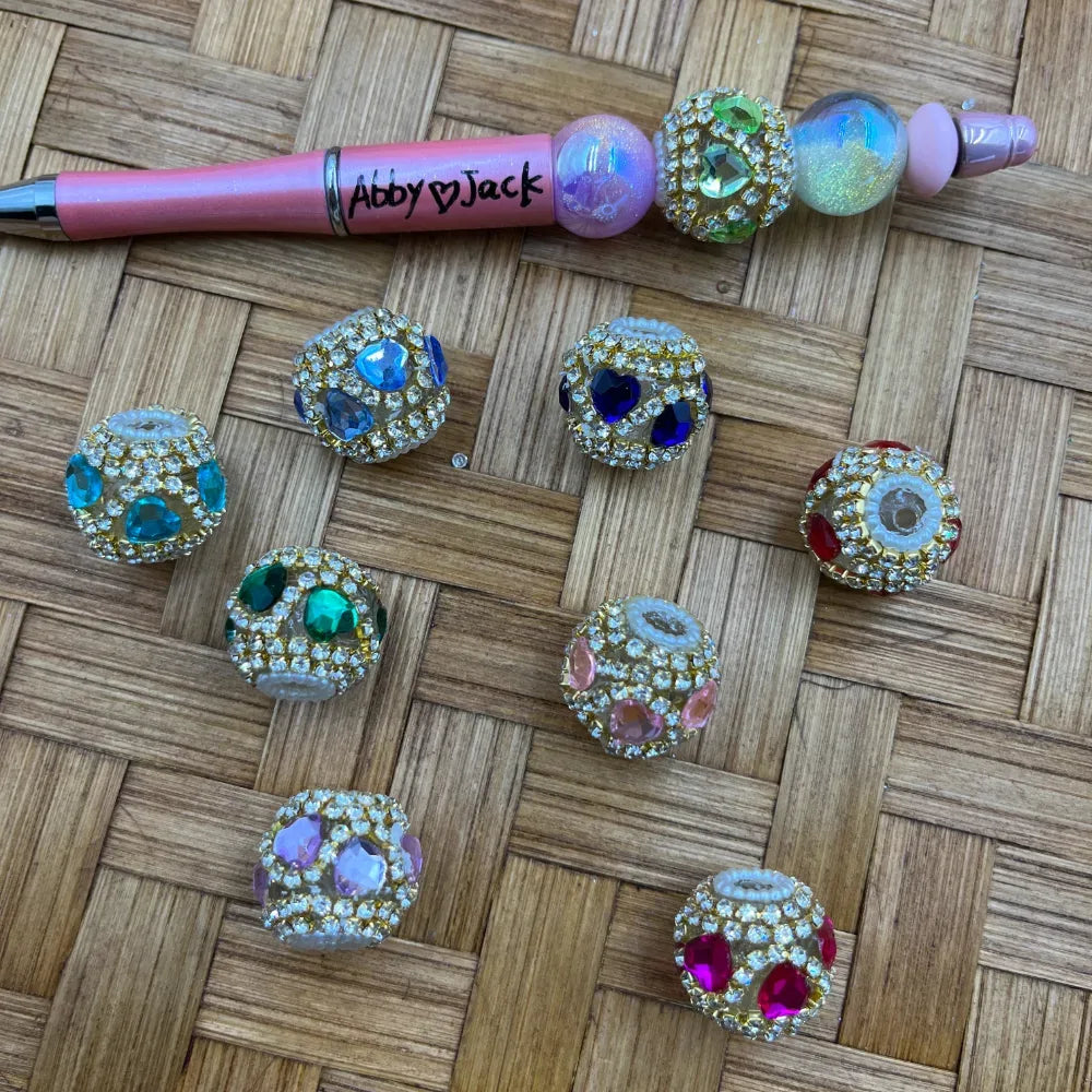 Assorted rhinestone pave fancy beads with colorful heart and gem accents in blue, green, pink, teal, and red, displayed on woven surface alongside a decorated beadable pen labeled Abby and Jack.