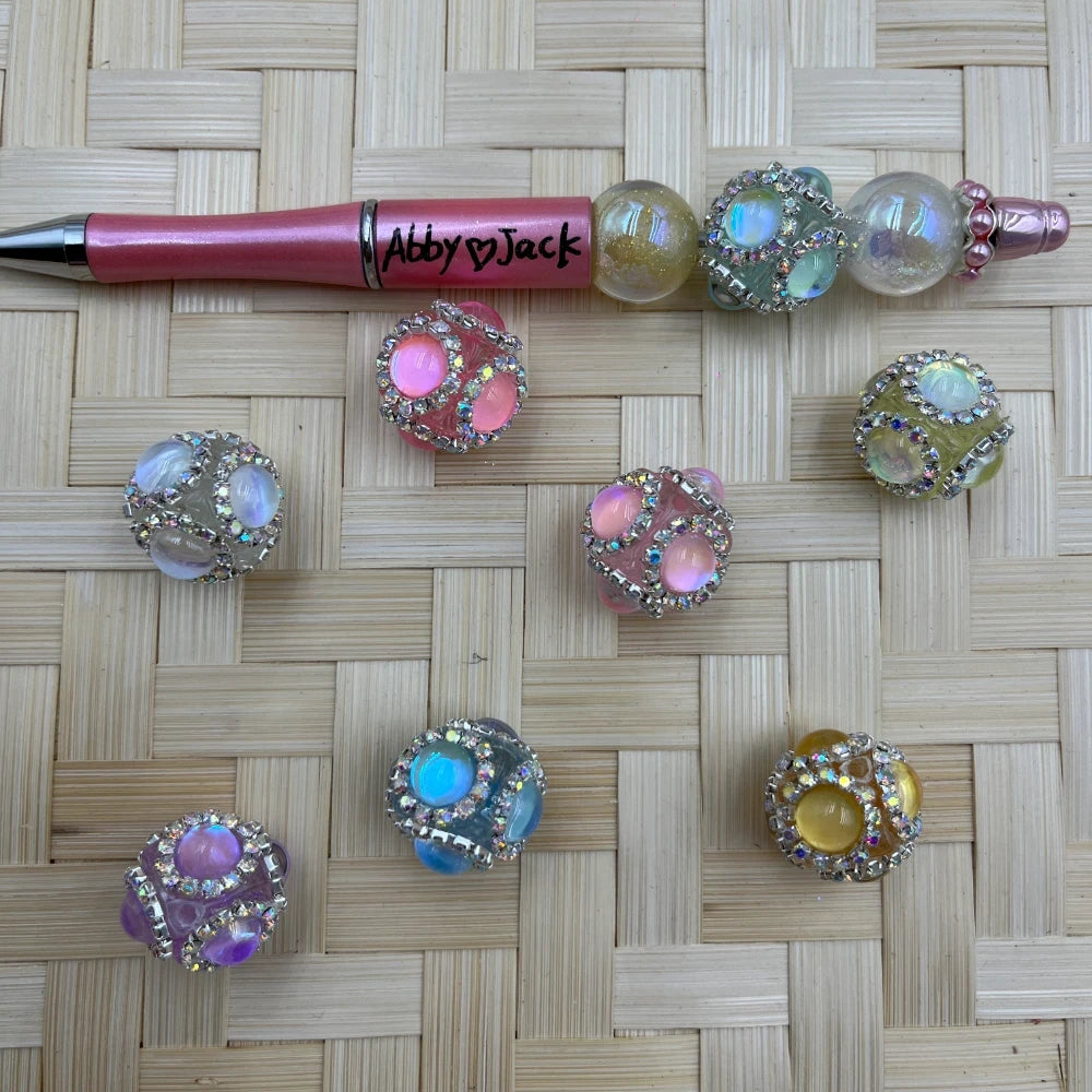 Assorted pastel rhinestone paw print bubblegum fancy beads in pink, purple, blue, yellow and clear iridescent colors displayed on woven surface next to a pink beadable pen labeled Abby Jack.