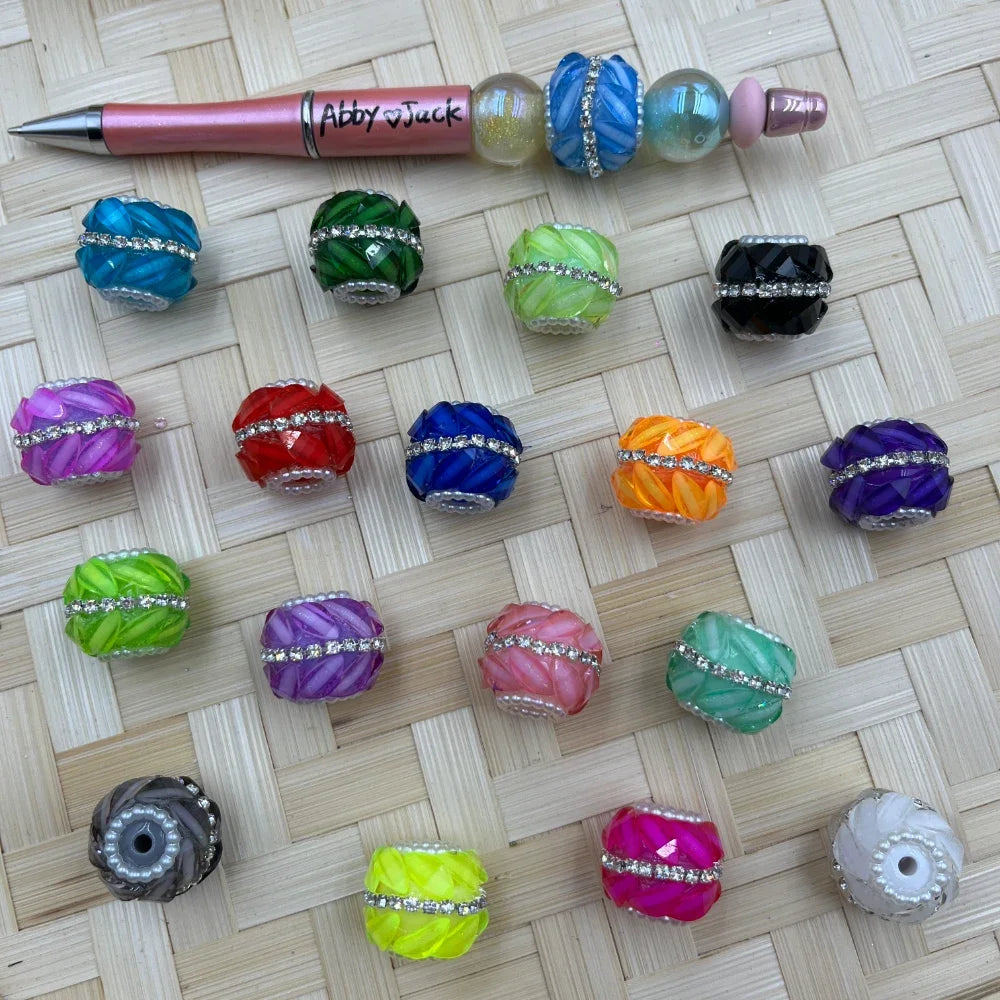 Assorted colorful faceted acrylic fancy beads with rhinestone and pearl bands in 20 colors displayed on woven bamboo surface next to a beadable pen.