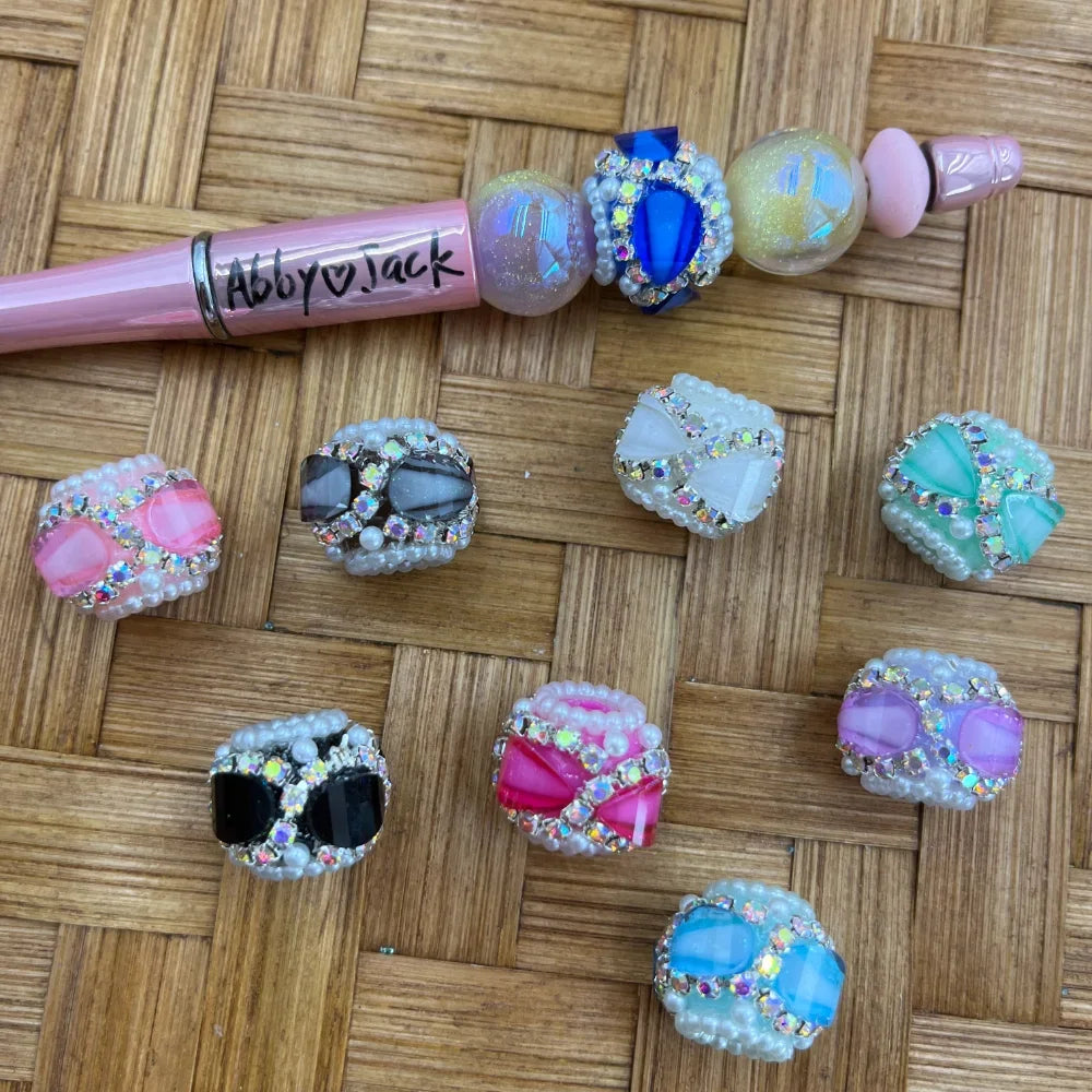 Colorful rhinestone and pearl-edged bow fancy beads in pink, black, white, mint, purple, blue, and hot pink displayed on a woven surface next to a decorated beadable pen labeled Abby and Jack.