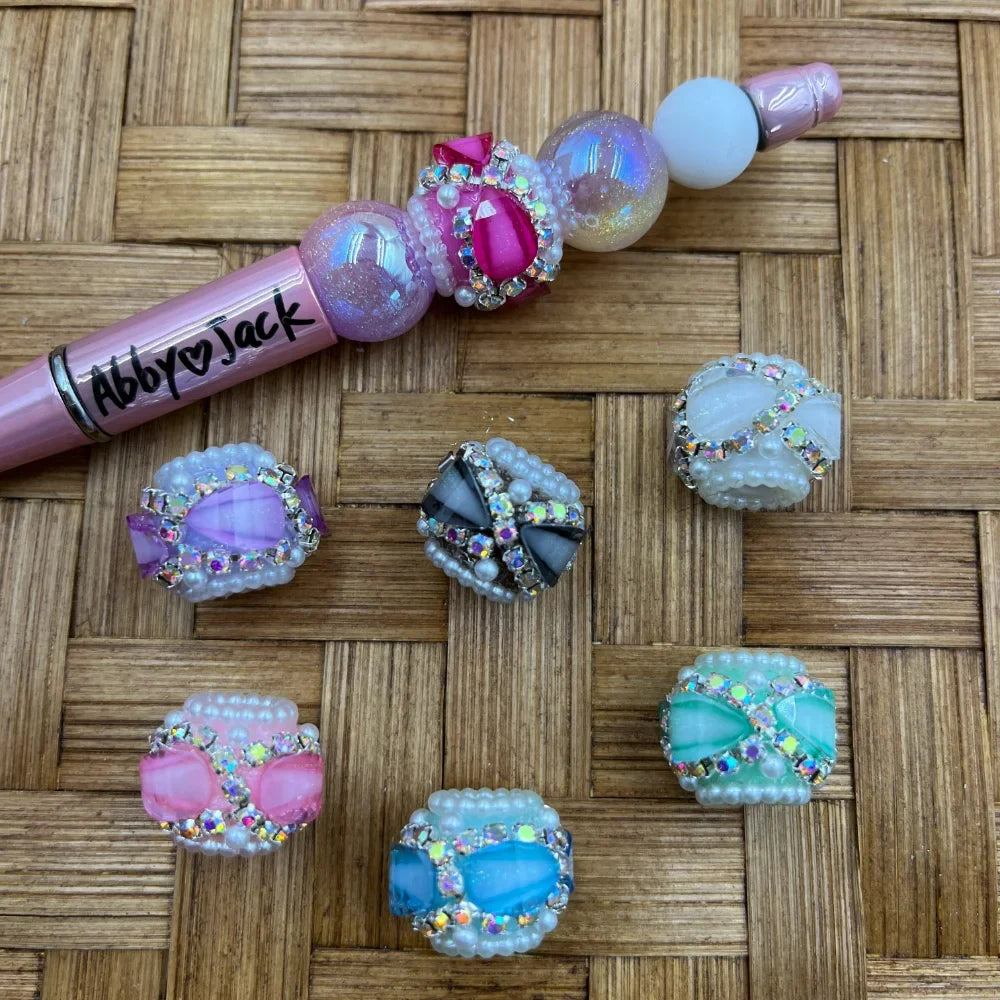 Assorted pastel rhinestone pearl bow fancy beads in purple, pink, gray, white, blue, and mint green colors displayed on a woven surface next to a decorated beadable pen branded Abby Jack.