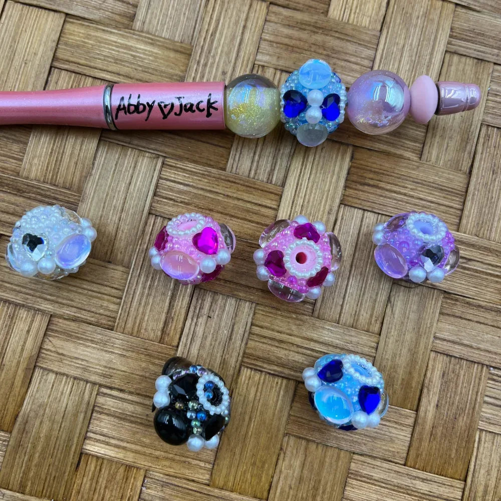 Assorted rhinestone and pearl cluster fancy beads in pink, blue, black, white, and purple with heart gem accents, displayed on a woven surface alongside a pink glitter beadable pen branded Abby Jack.