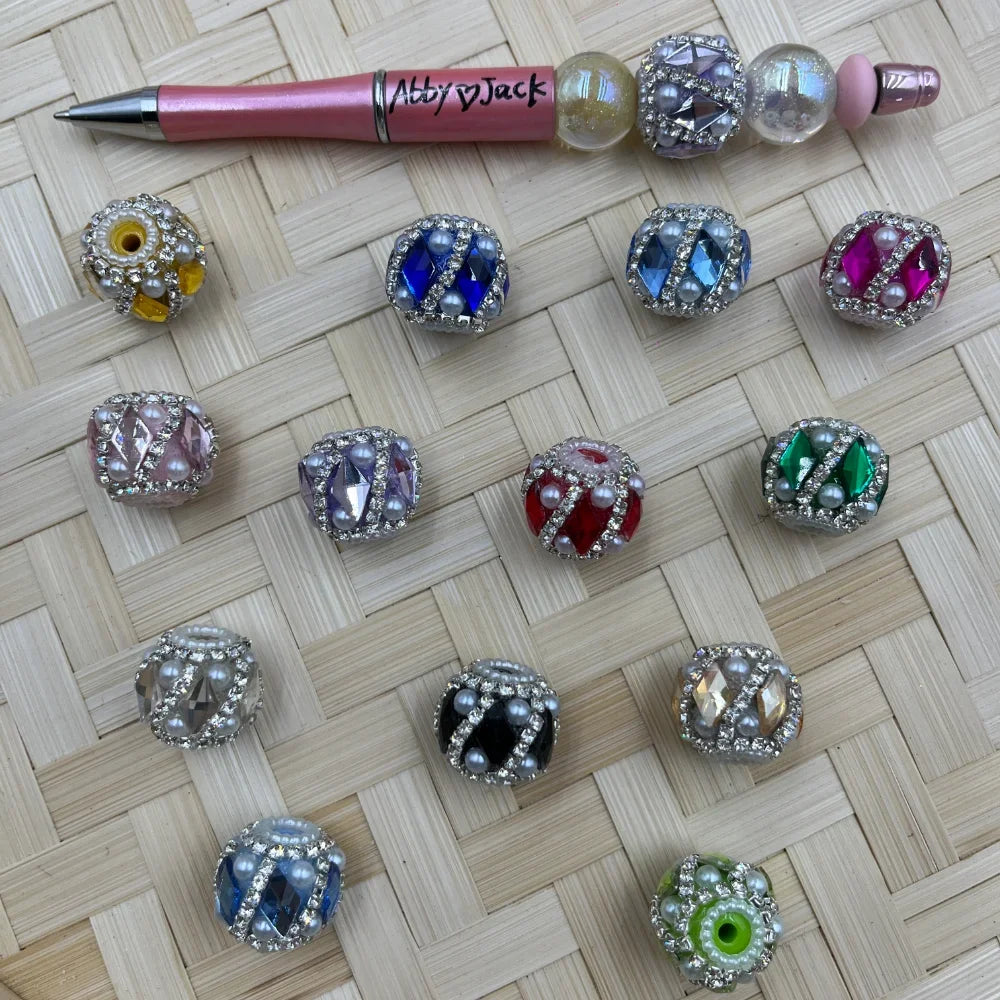 Assorted rhinestone and pearl crystal fancy beads in multiple colors including blue, red, green, pink, black, and champagne with silver rhinestone borders, shown next to a pink beadable pen on a wicker background.
