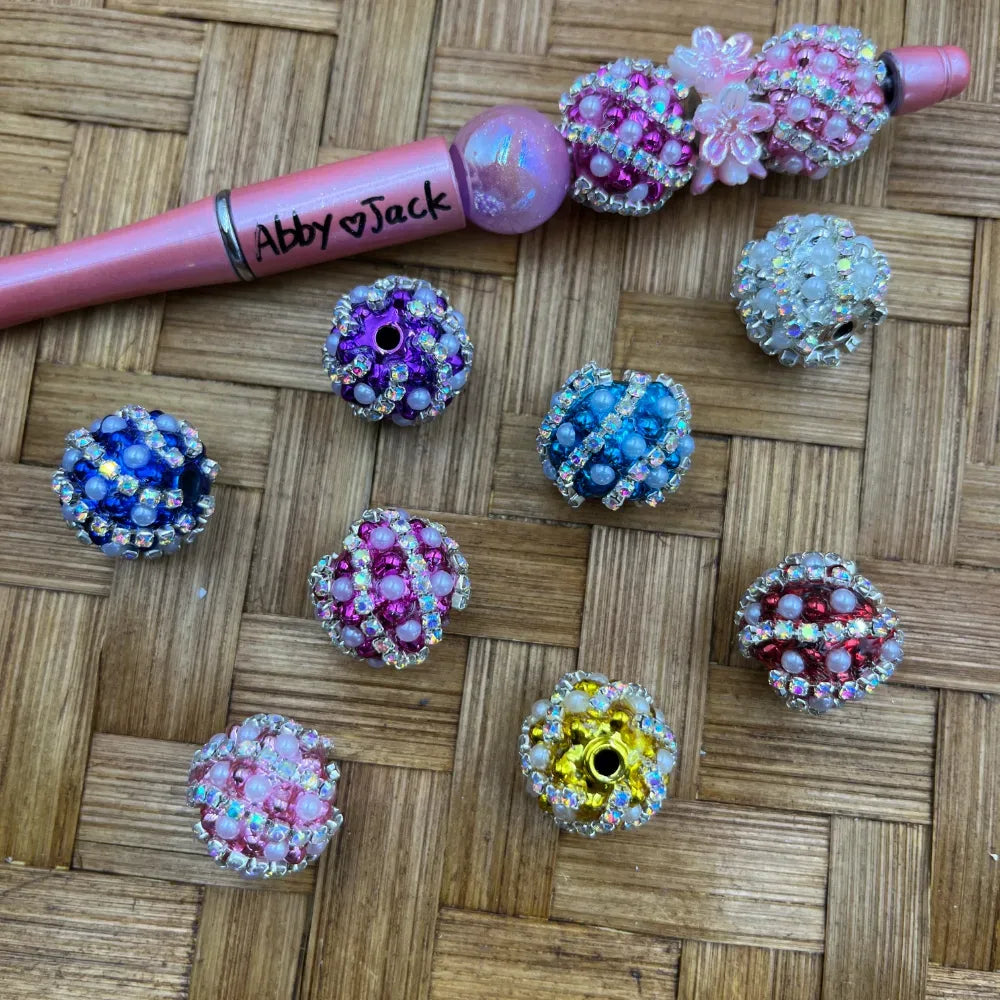 Assorted rhinestone and pearl fancy beads in 8 colors including blue, purple, pink, teal, red, yellow, and white, displayed on a woven surface next to a pink beadable pen by Abby Jack.