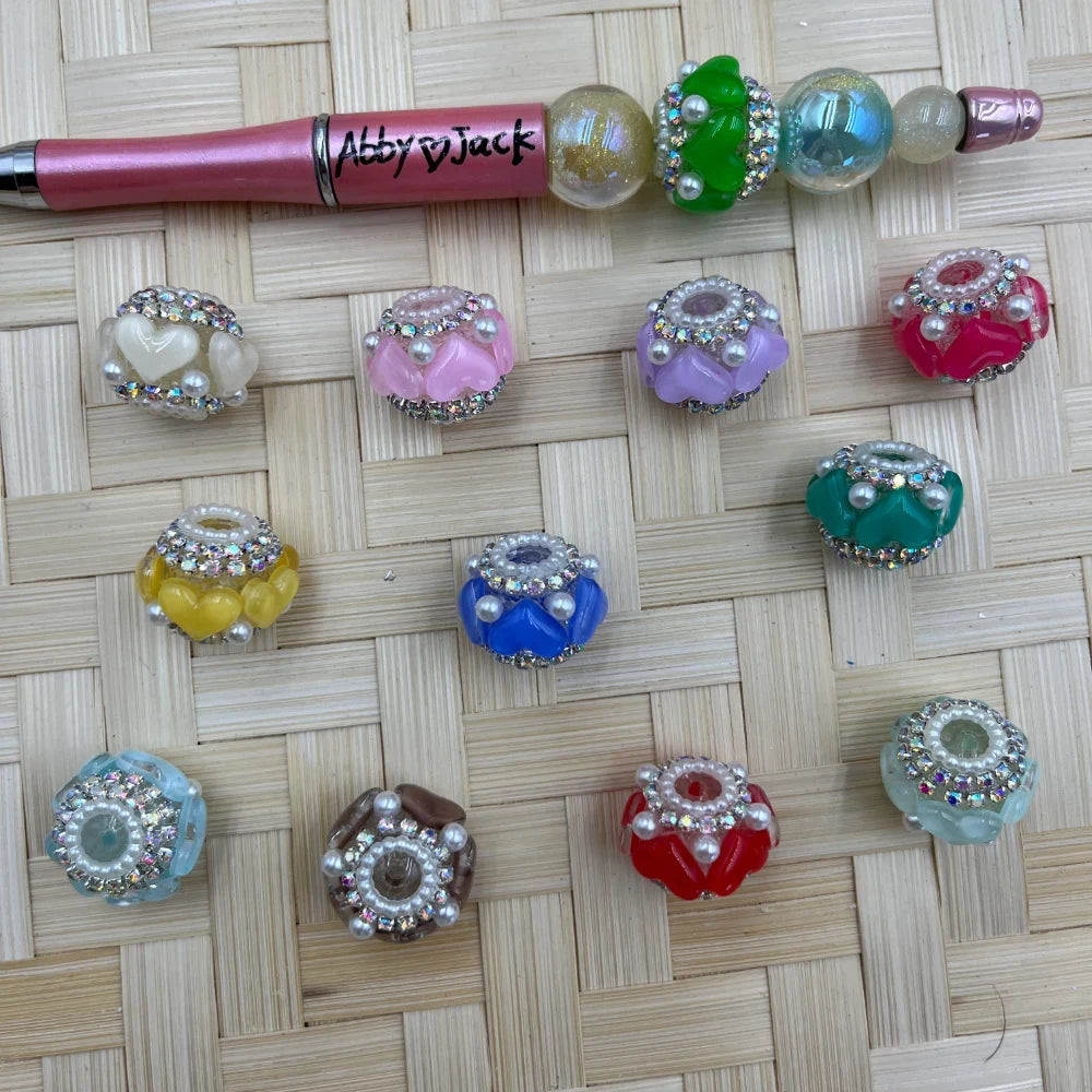 Assorted colorful rhinestone and pearl embellished fancy rondelle beads in pink, blue, red, green, teal, yellow, lavender, brown, and white displayed on a woven surface next to a beadable pen.