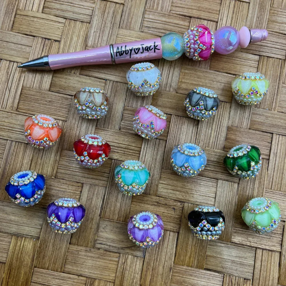 Assorted rhinestone and pearl trimmed crystal fancy beads in multiple colors including red, blue, green, purple, orange, teal, pink, black, and white, displayed next to a beaded pen on a wicker surface.