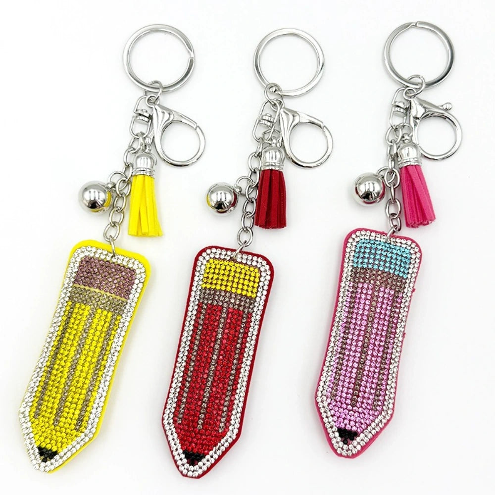 Three rhinestone-encrusted pencil keychain charms in yellow, red, and pink with matching tassels and silver bell dangles on key rings.